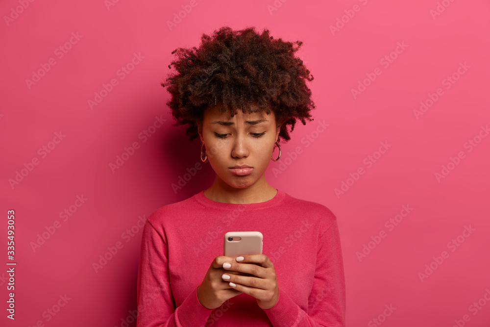 Distressed ethnic woman with sad unhappy expression, checks newsfeed on smartphone, purses lips and feels regret, expresses dissatisfaction as didnt get message from boyfriend, stands indoor alone Distressed ethnic woman with sad unhappy expression, checks newsfeed on smartphone, purses lips and feels regret, expresses dissatisfaction as didnt get message from boyfriend, stands indoor alone