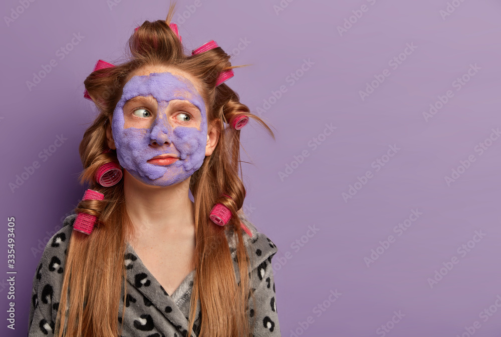 Serious small lady looks away, applies beauty face mask, wears hair curlers, prepares for special occasion for children, wears domestic robe, isolated on purple background, copy space for promo Serious small lady looks away, applies beauty face mask, wears hair curlers, prepares for special occasion for children, wears domestic robe, isolated on purple background, copy space for promo