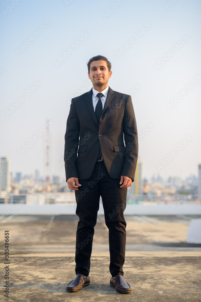 Full body shot of young Persian businessman in suit against view of the city Full body shot of young Persian businessman in suit against view of the city