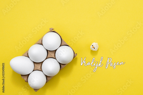Vrolijk Pasen, Dutch words for Happy Easter. An egg carton with six white eggs and a toy baby chicken on a yellow background. Room for text. 