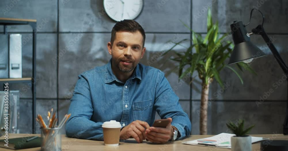 Happy handsome businessman tapping on smartphone while sitting at table at work and smiling to camera. Cheerful Caucasian male with beard texting on cellphone with smile on face. Office worker concept Happy handsome businessman tapping on smartphone while sitting at table at work and smiling to camera. Cheerful Caucasian male with beard texting on cellphone with smile on face. Office worker concept
