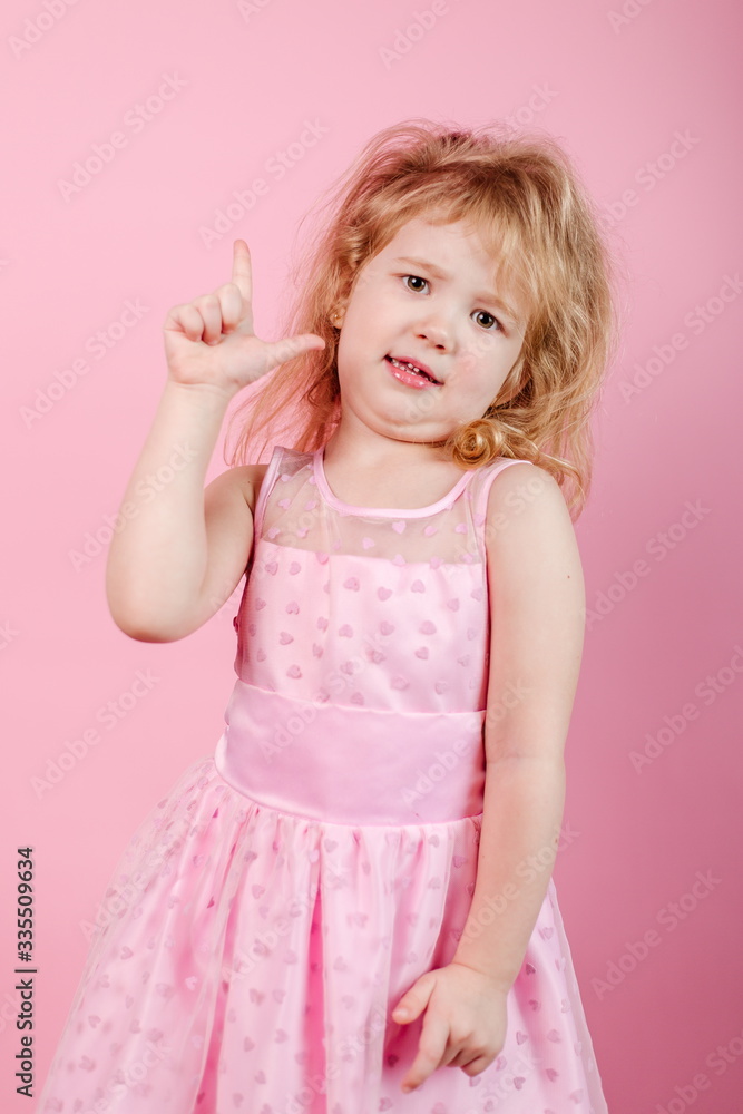 Little funny girl in a pink dress standing on a pink background showing thumb up