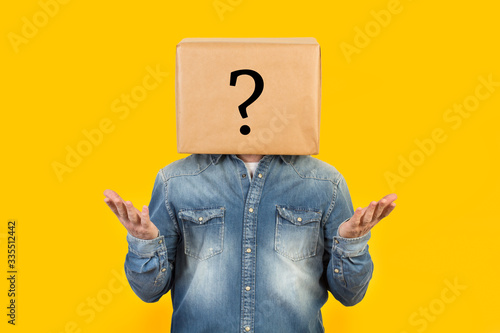 Hombre con caja de cartón en la cabeza con un signo de interrogación y gesto con las manos levantadas hacia arriba sobre fondo amarillo liso brillante aislado. Vista de frente. Copy space