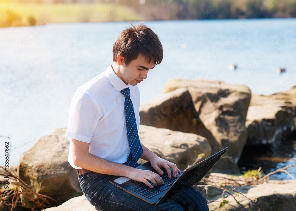  One Boy wearing white shirt, tie and jeans studying /learning online on the computer outdoors near the lake. View from the back