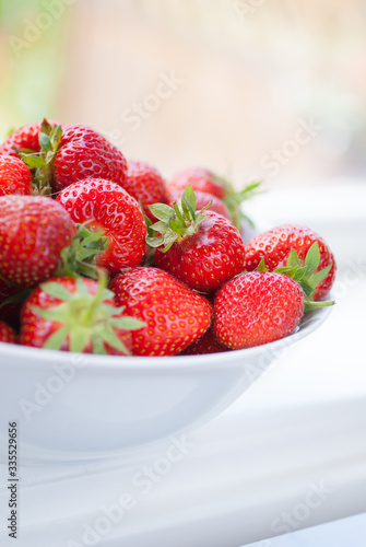 Pile of ripe  red strawberries in a white bowl on window sill. Blurred background. Bright  colourful  midsommar.