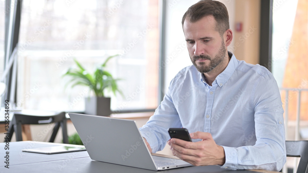 Young Man using Laptop and Smartphone in Office Young Man using Laptop and Smartphone in Office