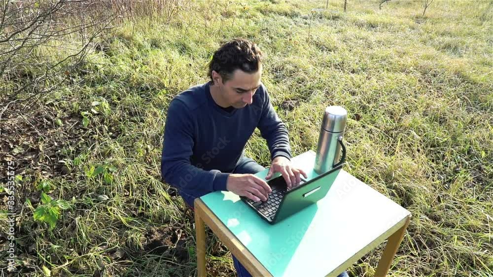 Young man developer working remote outdoors in yard, sitting with laptop and table in park, isolation concept Young man developer working remote outdoors in yard, sitting with laptop and table in park, isolation concept