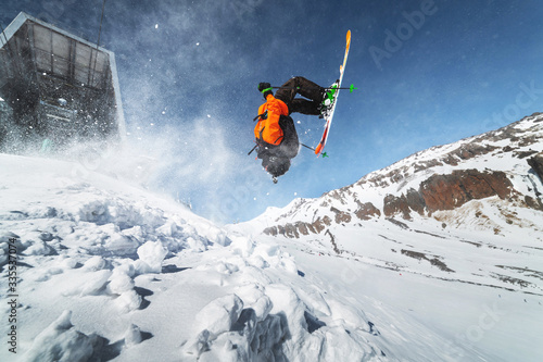 Low angle view athlete skier in an orange jacket does a back flip with flying powder of snow against a clear blue sky and snow-capped mountains of the Caucasus.
