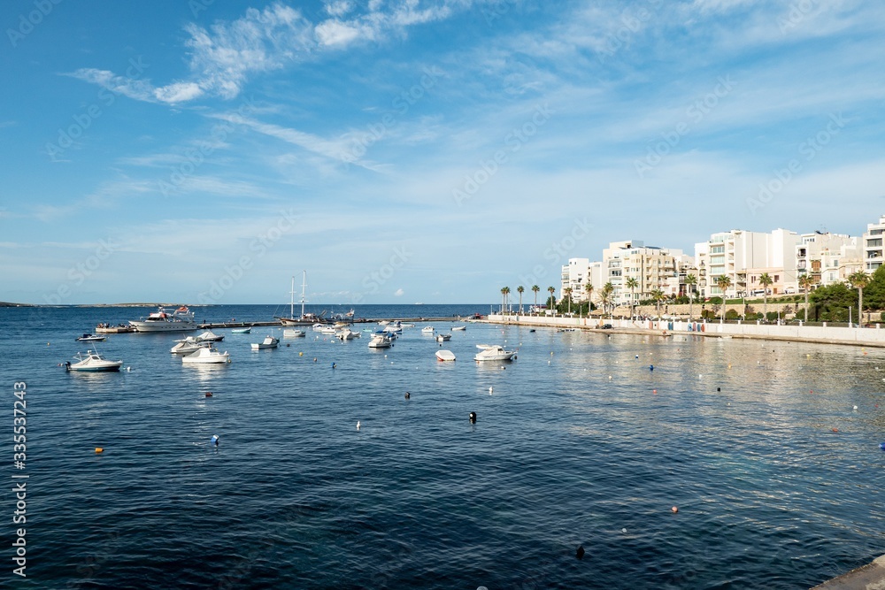 The cityscape of Bugibba city in Malta with a view at the sea, port, palms, apartments and the Dawret il-Gzejjer street reflected on water surface The cityscape of Bugibba city in Malta with a view at the sea, port, palms, apartments and the Dawret il-Gzejjer street reflected on water surface