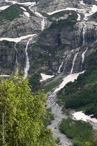 A multi-jet alpine waterfall on a high cliff with a green forest at the foot with glimpses of snow. The concept of a natural landscape in a natural environment on a cloudy day.