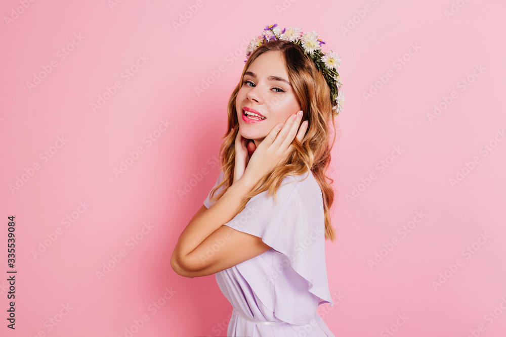 Graceful white lady touching her face and smiling. Indoor portrait of magnificent blonde girl in flower wreath standing on pink background.