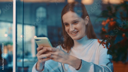 View through the window of beautiful happy european young woman using cellphone or smartphone at coffee shop, texing message, watching video or bloggs. Close up
