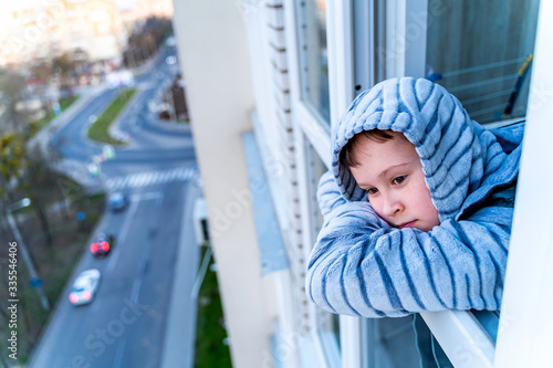 Quarantine. Little boy stands and looka out of a window bored. Longing for fresh air and walks along the street. Forced home during quarantine due to the coronavirus pandemic. photo