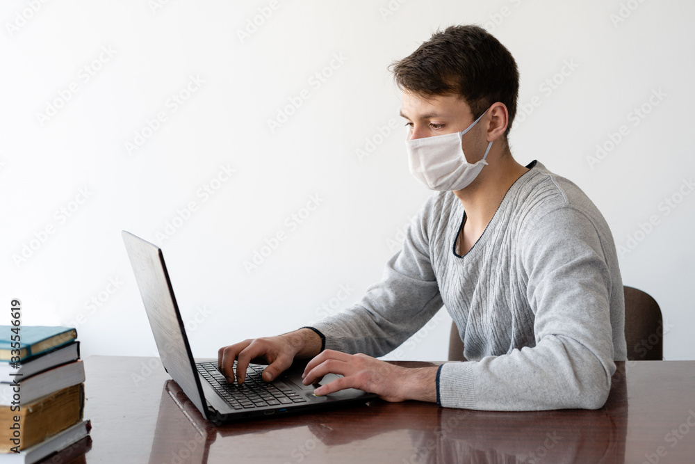 A young man with a medical face shield works on his laptop at home. Learning and working online at home during quarantine during the coronavirus pandemic. A young man with a medical face shield works on his laptop at home. Learning and working online at home during quarantine during the coronavirus pandemic.