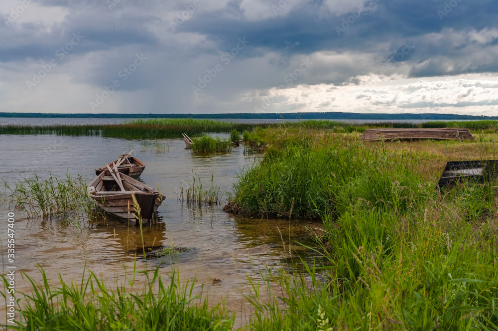 Wooden fishing boats are empty on a lake in a thicket of grass. Thunderclouds over the lake in the fishing season. Empty wooden old boats on the lake. Nature, landscape, lakes in Belarus.