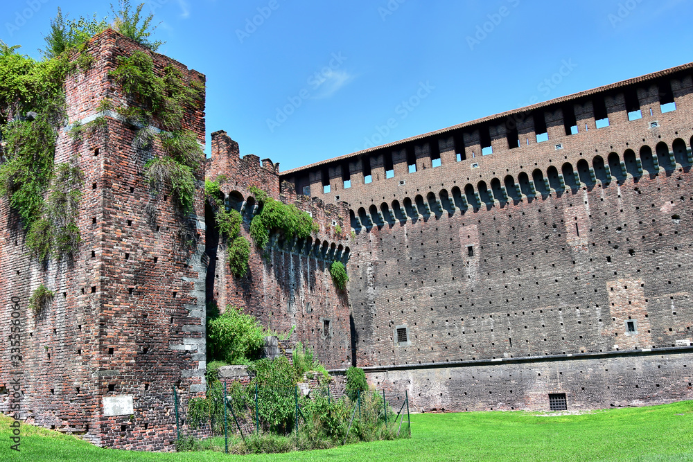 Milan, Italy - Castello Sforzesco or Sforza Castle, walls of the fortress made of tiled bricks, covered with green plants, grass in front of the castle, blue sky.
