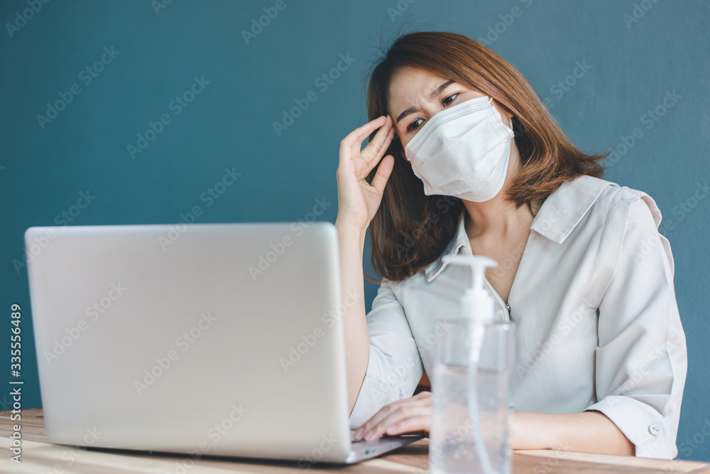 A beautiful businessman in a white dress with a mask and starts to get headache while working with a laptop in the office Alcohol gel placed on her desk A beautiful businessman in a white dress with a mask and starts to get headache while working with a laptop in the office Alcohol gel placed on her desk
