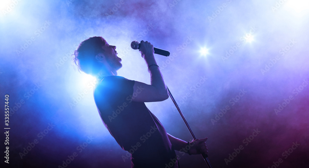 Young male singer performing on a stage and playing the guitar. Smoky blue background - obrazy, fototapety, plakaty 