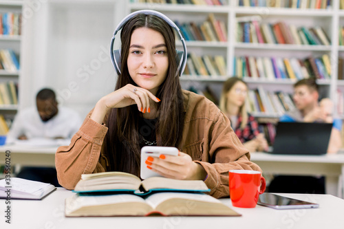Close up portrait of charming cute smiling young girl student, sitting in the library at the table with many books, listening music in earphones, enjoying the pause before study