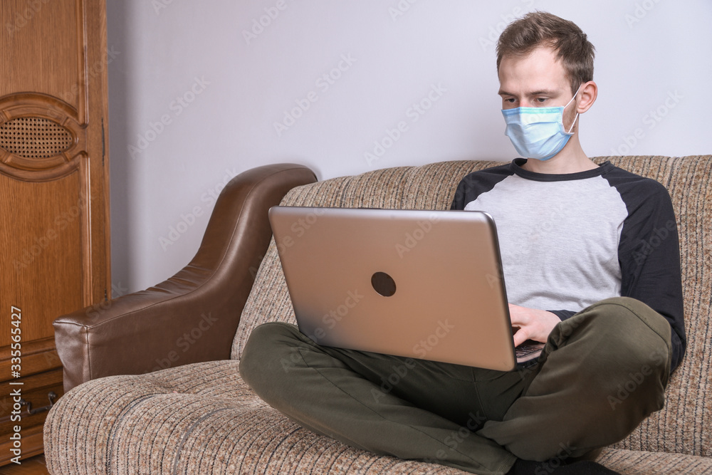 Young man in medical mask working at home in the room on the sofa using a laptop. Quarantine, self-isolation, coronavirus protection. Vacation from work. Young man in medical mask working at home in the room on the sofa using a laptop. Quarantine, self-isolation, coronavirus protection. Vacation from work.