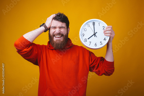 The time is so fast. Photo of bearded guy with eyes closed and hand on his head is holding a white clock.