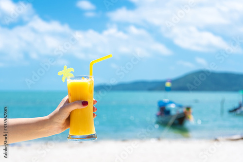 First-person view. Girl holds a glass cup of cold mango fresh on the background of a sandy tropical beach. White sand and a boat. Fairytale vacation in Thailand