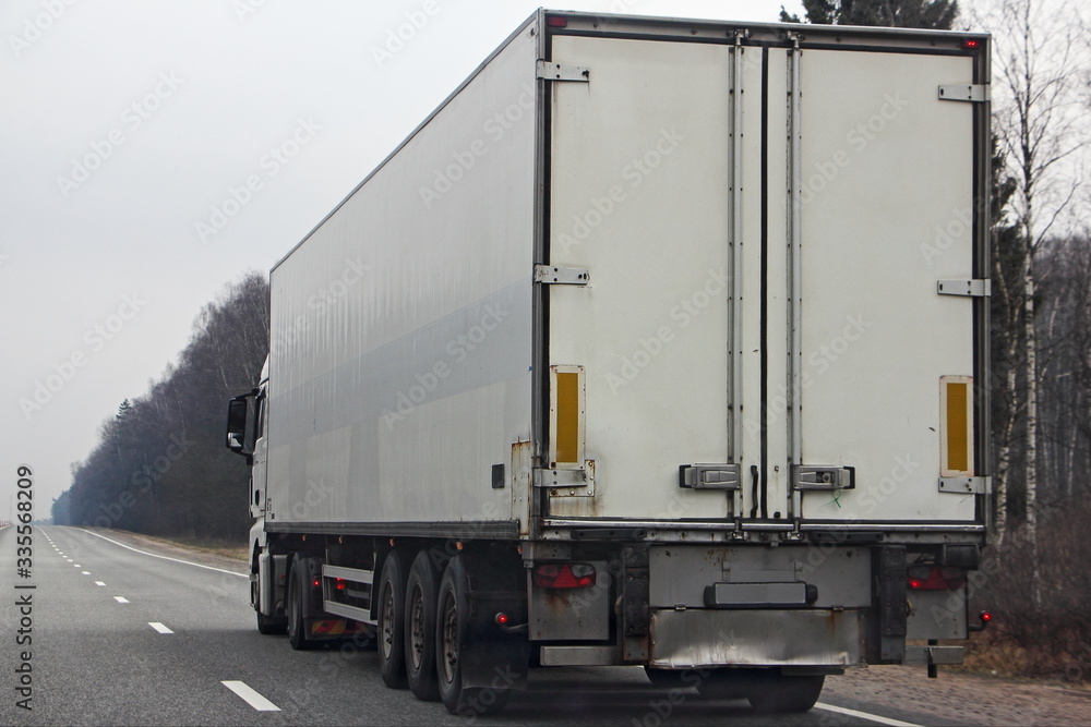 White semi truck close up with copy space blank on three-axle trailer rear door on empty highway road at spring day
