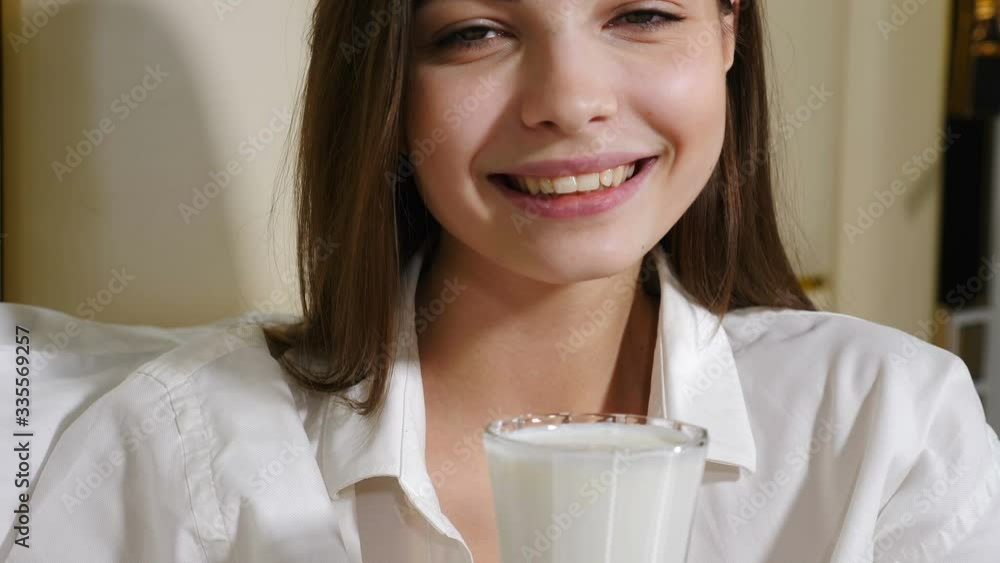 Portrait of attractive young woman smiling at camera drinking milk or other milk beverage. Beautiful girl on bed with white pillowsin on background. Breakfast in bed in morning. Shot in 4k Portrait of attractive young woman smiling at camera drinking milk or other milk beverage. Beautiful girl on bed with white pillowsin on background. Breakfast in bed in morning. Shot in 4k
