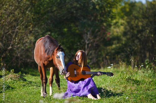 red-haired girl in a vintage purple dress plays the guitar next to the horses