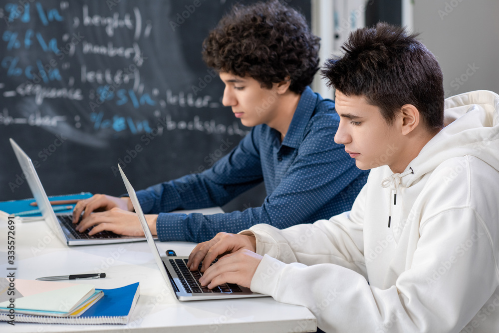 Two teenage guys typing in front of laptops while sitting by desk at lesson Two teenage guys typing in front of laptops while sitting by desk at lesson