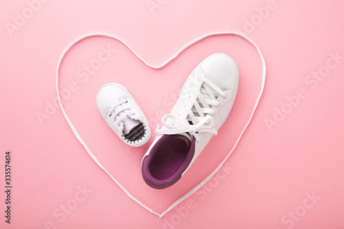 Mother and little kid shoes. Heart created from white shoelaces. Light pink floor background. Pastel color. Love sport together concept. Closeup. Top down view.