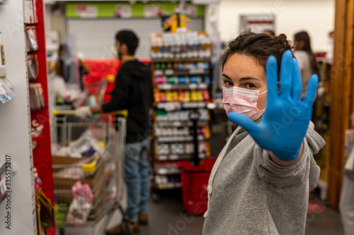 The girl in the supermarket in a protective mask and disposable gloves shows a hand symbol, stop sign. Look straight at the camera. The concept of protection and disinfection in times of viruses.