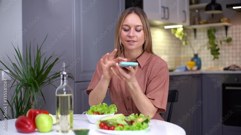Attractive young woman uses smartphone for taking photos of vegetables lying on kitchen table. Concept of social networks. Female taking photos of healthy vegetarian food on cell smart phone. Attractive young woman uses smartphone for taking photos of vegetables lying on kitchen table. Concept of social networks. Female taking photos of healthy vegetarian food on cell smart phone.