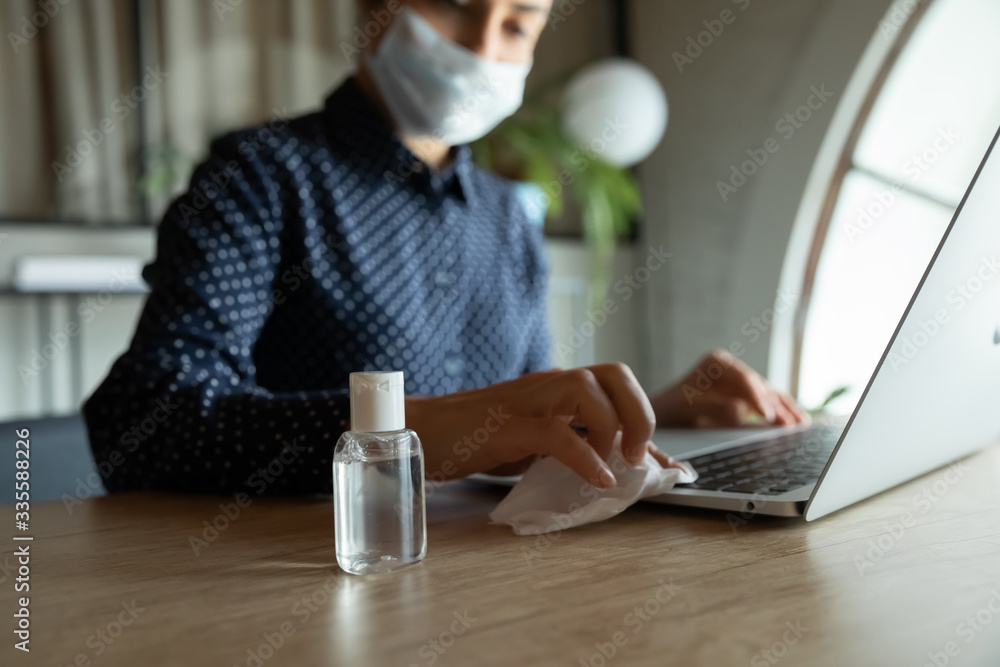 Focus on plastic bottle with antiseptic alcohol-based liquid standing on work desk with cautious woman in medical facemask sanitizing computer keyboard on background, stop spreading virus infection. Focus on plastic bottle with antiseptic alcohol-based liquid standing on work desk with cautious woman in medical facemask sanitizing computer keyboard on background, stop spreading virus infection.