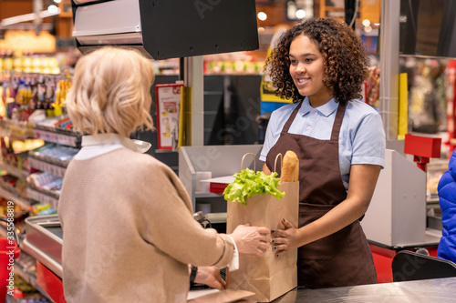 Pretty young cashier giving mature female paperbag with bread and groceries