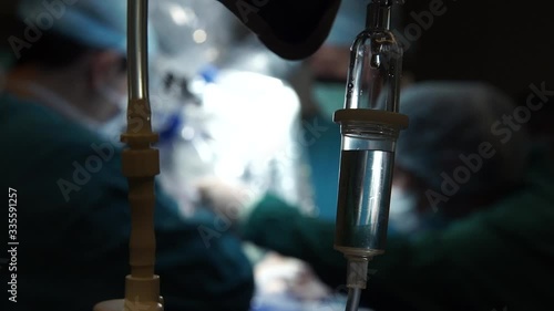 IV (infusion) bags and bottles hanging on poles during a real surgery going on in the background. Patient, illness, treatment, anesthesia, hospital, medicine and healthcare symbolism and concept.