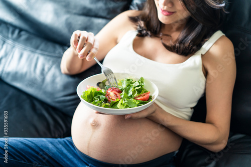 Pregnant women sitting on sofa is holding salad bowl in her left hand. She is holding fork with red tomato on right hand. Seen in top side view and focus on tomato. Healthy for mother concept. © Kawee