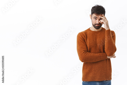Portrait of awkward and embarrassed bearded man pretend he not see his ex, hiding face behind hand, facepalm and peek through fingers, avoiding eye contact, standing white background