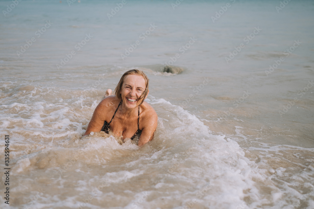 Happy smiling excited elderly senior woman tourist playing in water and swimming in the big waves on the ocean sea beach. Traveling along Asia, active lifestyle concept. Happy smiling excited elderly senior woman tourist playing in water and swimming in the big waves on the ocean sea beach. Traveling along Asia, active lifestyle concept.