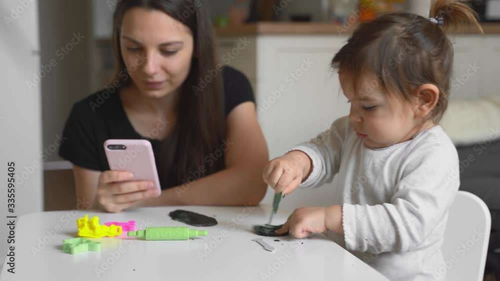 YOung mother working from home form phone with small toddler playing near mum. Home office, quarantine Covid-19 YOung mother working from home form phone with small toddler playing near mum. Home office, quarantine Covid-19