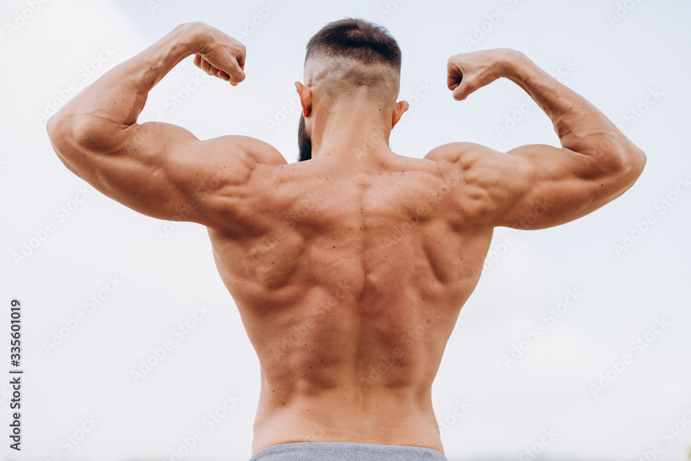 Handsome bearded bodybuilder man with perfect muscular body posing outdoors on the sky background. Strong man with naked torso standing, looking away. View from the back Handsome bearded bodybuilder man with perfect muscular body posing outdoors on the sky background. Strong man with naked torso standing, looking away. View from the back