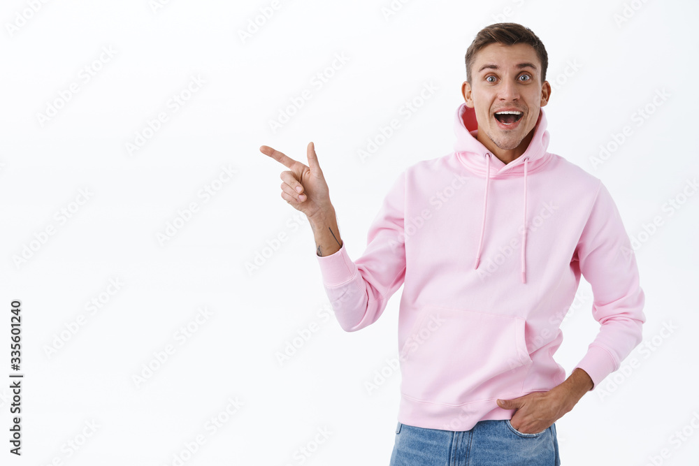 Cheerful upbeat good-looking blond man, wearing pink hoodie react joyful to wonderful news, look camera with beaming smile excited, showing way, pointing finger left, white background Cheerful upbeat good-looking blond man, wearing pink hoodie react joyful to wonderful news, look camera with beaming smile excited, showing way, pointing finger left, white background