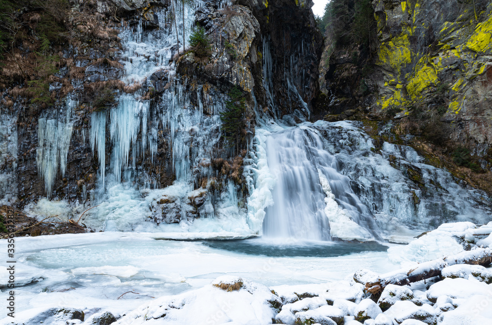 The Reinbach waterfall in Valle Aurina (Ahrntal), South Tyrol, Italy. White stalactites come down from the rocks, the water quickly pours into the small frozen lake. Granite and light green lichens. The Reinbach waterfall in Valle Aurina (Ahrntal), South Tyrol, Italy. White stalactites come down from the rocks, the water quickly pours into the small frozen lake. Granite and light green lichens.