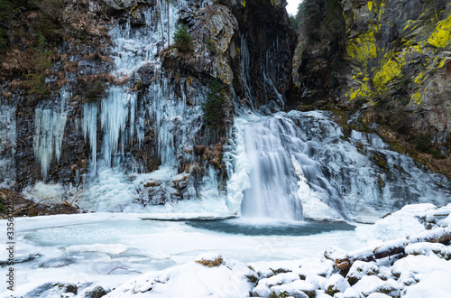 The Reinbach waterfall in Valle Aurina (Ahrntal), South Tyrol, Italy. White stalactites come down from the rocks, the water quickly pours into the small frozen lake. Granite and light green lichens.