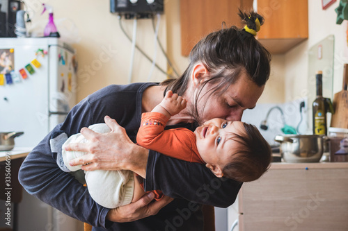 Hipster young father hug and kiss his baby infant boy holding on hands. At home on kitchen background. Lifestyle photo of family care and happiness concept. Middle class. 