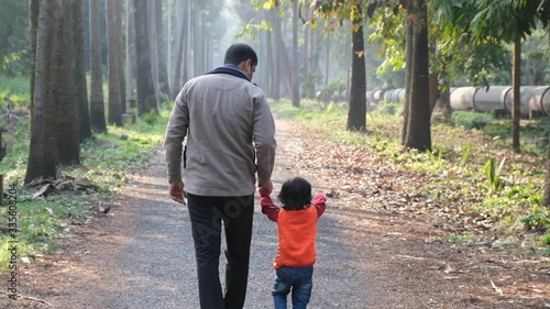 Backside of an Indian brunette father and his baby boy in winter garments walking in a forest path in winter afternoon in natural green background. Indian lifestyle and parenthood.