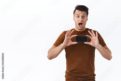 Wow cool, check it out. Surprised and wondered handsome adult man in brown t-shirt react at new video went viral on smartphone app, showing mobile phone display horizontally, recommend link