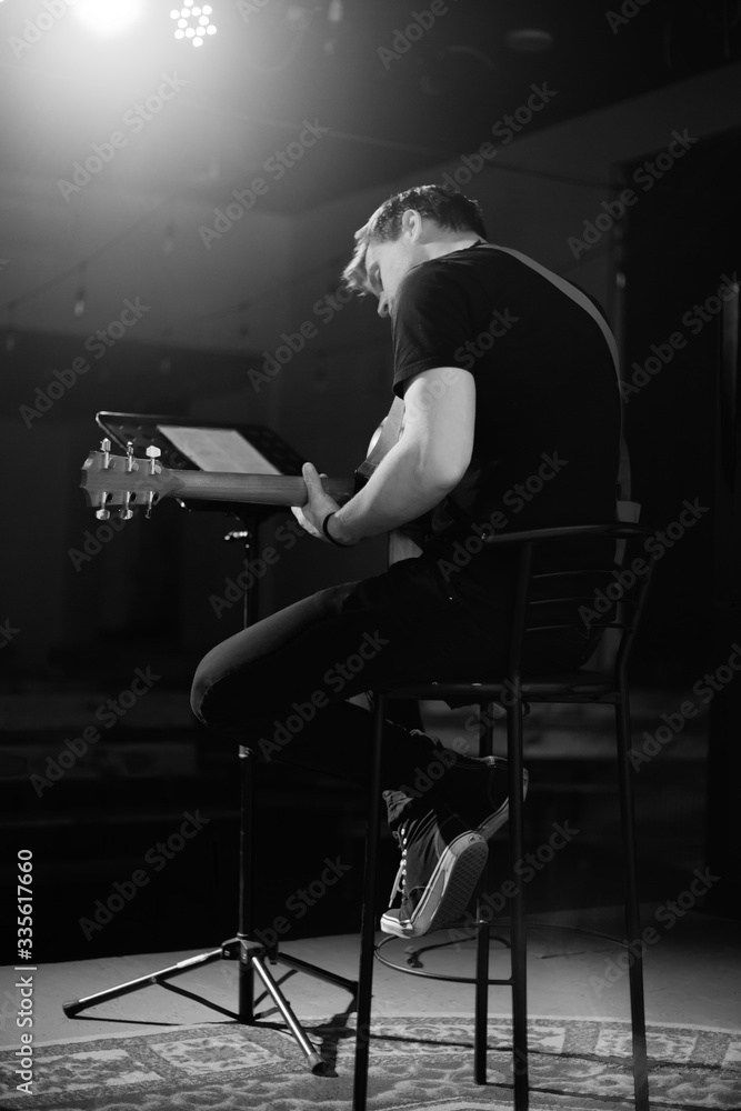 A young handsome guy of European appearance plays an acoustic guitar and sits on a chair. Black and white photography. A young handsome guy of European appearance plays an acoustic guitar and sits on a chair. Black and white photography.