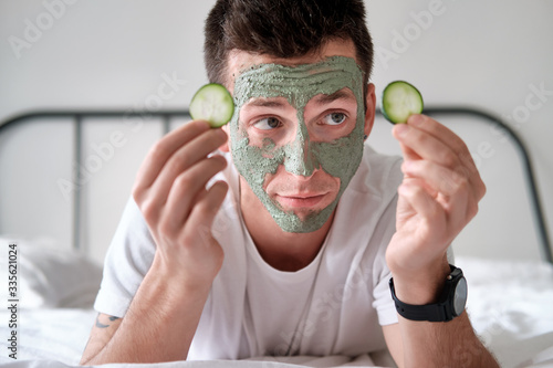 Young man in a white shirt with applied green cosmetic mask holding pieces of cucumber