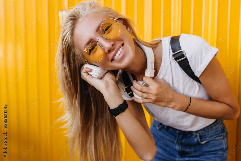Fototapeta premium Wonderful girl in white t-shirt and jeans expressing positive emotions. Refined lady listening music in headphones on yellow background.